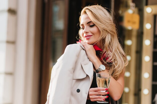 Prachtige jonge vrouw met elegant kapsel wegkijken en glimlachend staande. Outdoor portret van geïnspireerde blonde dame met rode manicure met wijnglas.