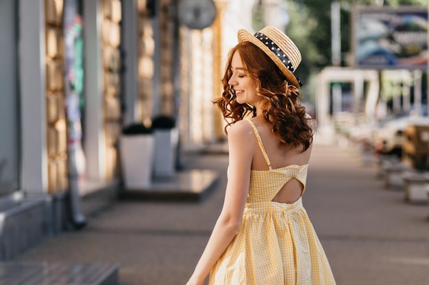 Prachtige roodharige meisje in vintage gele jurk poseren op stad achtergrond Outdoor foto van achterkant mooie vrouw met krullend kapsel chillen in zomerstad