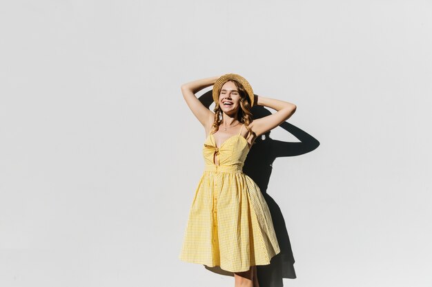 Prachtige slanke dame in hoed poseren in zonnige dag en lachen. Buiten schot van goedgehumeurd vrouwelijk model in gele jurk.