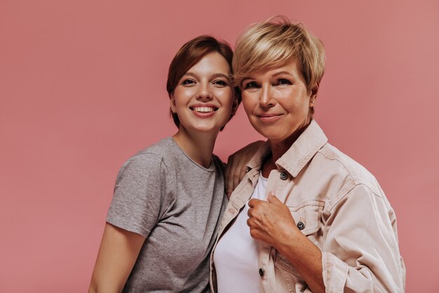 Prachtige twee vrouwen met moderne korte haarstijl in stijlvolle kleding glimlachen, knuffelen en kijken naar camera op roze achtergrond.