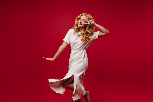 Prachtige vrouw in witte jurk die geluk uitdrukt. Aanbiddelijk blondemeisje die op rode muur dansen.