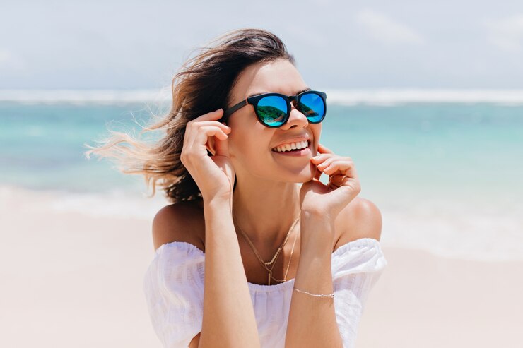 Gratis Foto prachtige vrouw in witte kledij en sprankelende bril poseren met blij gezicht expressie in hete zomerdag. aangename kaukasische vrouw die zich dichtbij oceaan op hemel bevindt