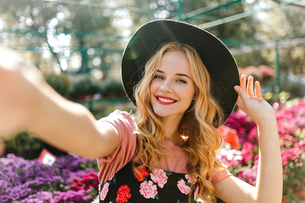 Gratis foto prachtige vrouw met mooie ogen selfie maken op de oranjerie. tevreden vrouw in zwarte hoed poseren met bloemen.