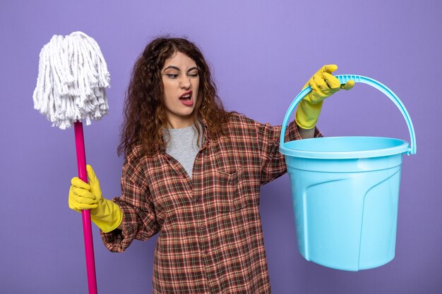 Prettige jonge schoonmaakster met handschoenen die een dweil vasthoudt en naar een emmer in haar hand kijkt, geïsoleerd op een paarse muur