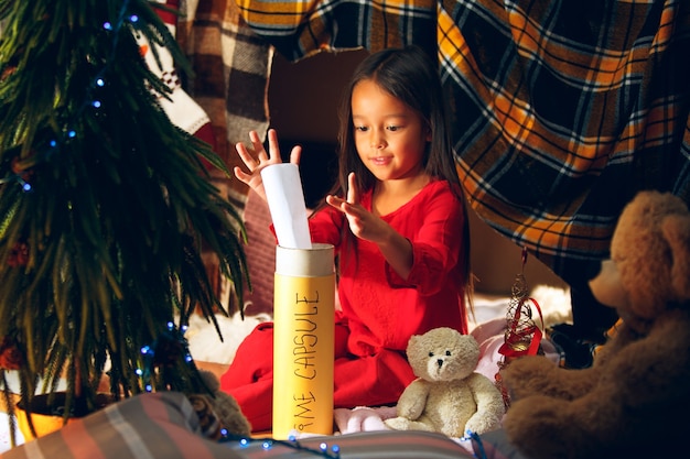 Gratis foto prettige kerstdagen en fijne feestdagen. schattig klein kind meisje schrijft de brief aan de kerstman in de buurt van de kerstboom