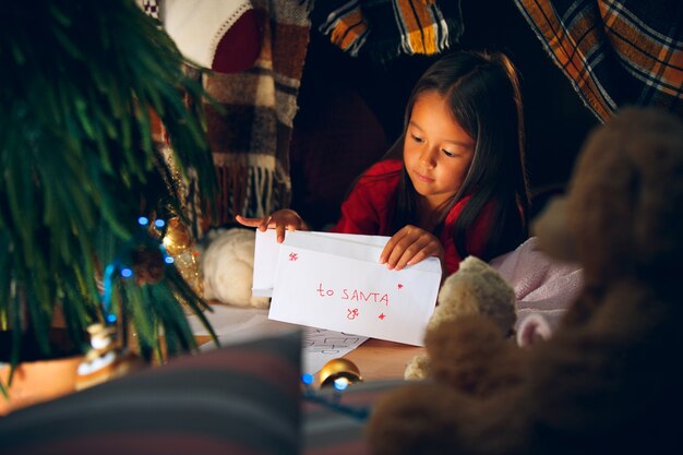 Prettige kerstdagen en fijne feestdagen. Schattig klein kind meisje schrijft de brief aan de kerstman in de buurt van de kerstboom