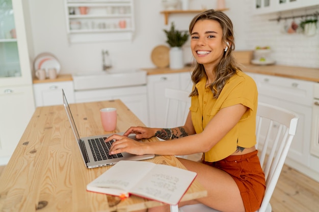 Pretty miling student vrouw met behulp van laptop en het schrijven van notities over haar moderne lichte keuken