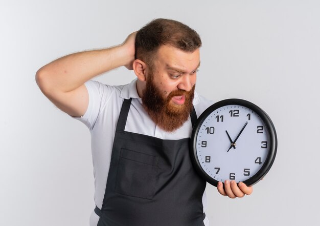 Professionele bebaarde kapper man in schort houden klok kijken naar het kijken verward en verrast staande over witte muur