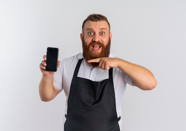 Professionele bebaarde kapper man in schort met smartphone wijzend met de vinger naar het glimlachend blij en opgewonden staande over witte muur