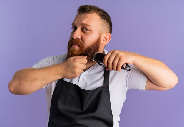 Professionele bebaarde kapper man in schort zijn baard trimmen met scheermachine op zoek verward staande over paarse muur