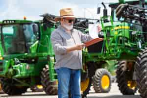 Gratis foto professionele boer met een moderne tractor aan het werk met documenten. ziet er zonnig uit. landbouw, tentoonstelling, machines, plantaardige productie. senior man in de buurt van zijn machine.