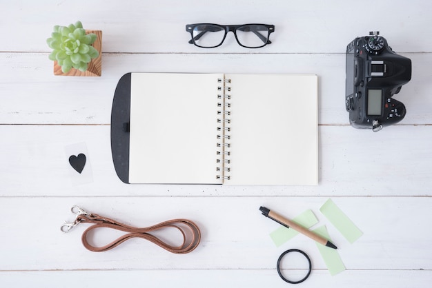 Professionele camera; lege spiraalvormige blocnote; schouwspel; plakbriefjes; riem; pen; heartshape en vetplant op witte houten tafel