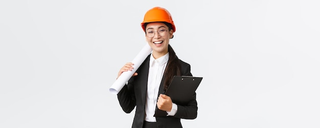Professionele gelukkige aziatische vrouwelijke architect bouwingenieur in helm en pak met blauwdrukken en klembord met bouwdocumentatie glimlachend vrolijke witte achtergrond