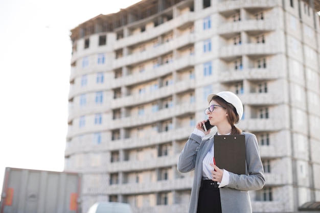 Gratis foto professionele jonge vrouwelijke architect die op het klembord van de cellphoneholding bij bouwwerf spreekt