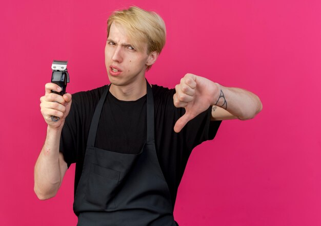 Professionele kapper man in schort bedrijf trimmer duimen omlaag wordt ontevreden weergegeven