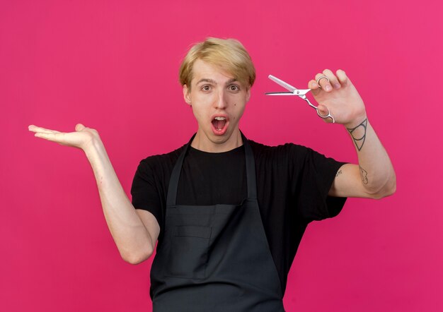 Professionele kapper man in schort houden schaar iets presenteren met arm van zijn hand wordt verrast en verbaasd