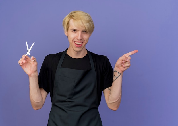 Professionele kapper man in schort houden schaar pointin met wijsvinger naar de kant lachend met blij gezicht