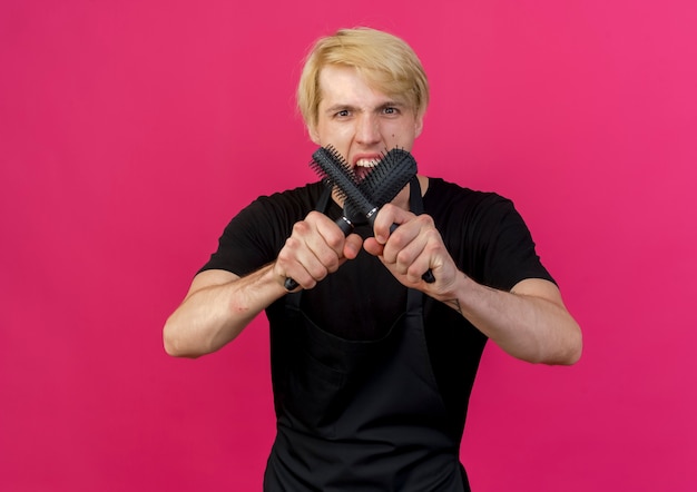 Professionele kapper man in schort met haarborstels schreeuwen met een boos gezicht