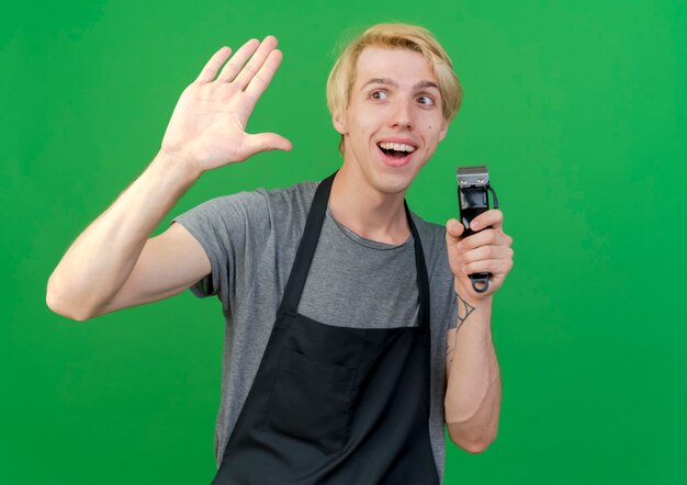 Professionele kapper man in schort met trimmer zwaaien met hand glimlachend vriendelijk staande over groene muur