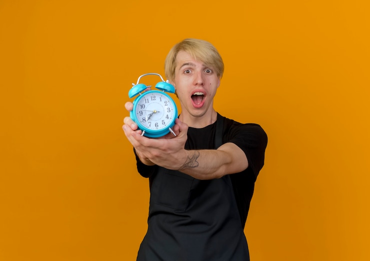 Gratis Foto professionele kapper man in schort tonen wekker emotioneel verbaasd en verrast staande over oranje muur