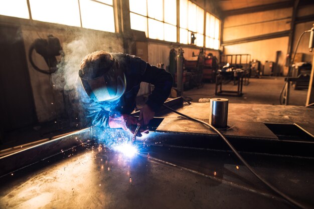 Professionele lasser in beschermende uniform en helm lassen metalen onderdeel in werkplaats