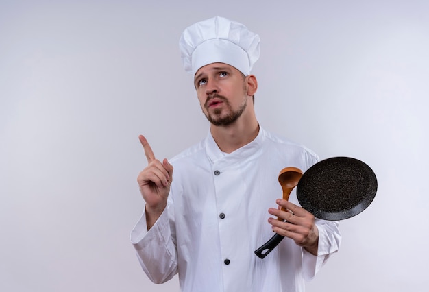 Professionele mannelijke chef-kok in wit uniform en kok hoed bedrijf koekenpan en houten lepel opzoeken wijzende vinger met peinzende uitdrukking staande op witte achtergrond