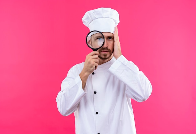 Professionele mannelijke chef-kok in wit uniform en kok hoed camera kijken door vergrootglas staande over roze achtergrond