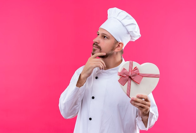 Professionele mannelijke chef-kok in wit uniform en kok hoed houden geschenkdoos in een hartvorm opzij kijken met hand op kin met peinzende uitdrukking op gezicht staande over roze achtergrond