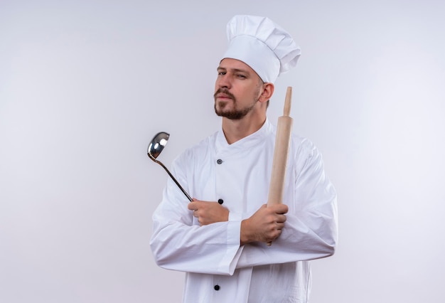 Professionele mannelijke chef-kok in wit uniform en kok hoed houden pollepel en deegroller kijken camera met verdachte uitdrukking staande op witte achtergrond