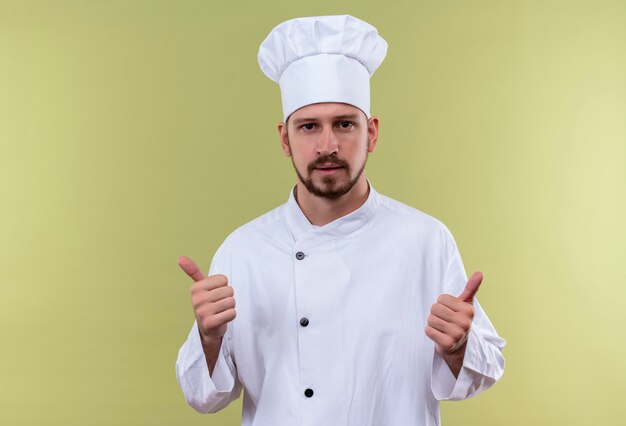 Professionele mannelijke chef-kok in wit uniform en kok hoed kijken camera duimen opdagen staande over gree achtergrond