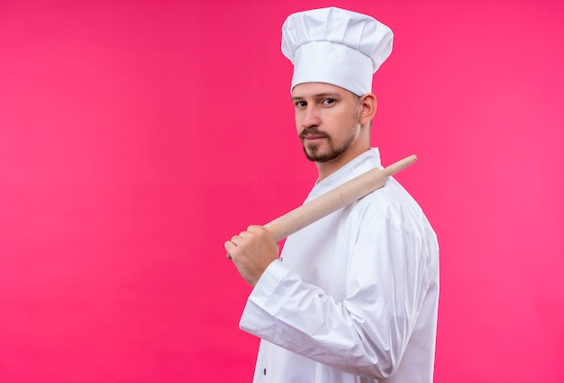 Professionele mannelijke chef-kok in wit uniform en kok hoed met rollende roze kijken zelfverzekerd staande over roze achtergrond