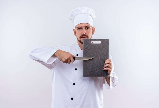 Professionele mannelijke chef-kok in wit uniform en koken hoed met snijplank en mes op zoek naar vertrouwen staande op witte achtergrond