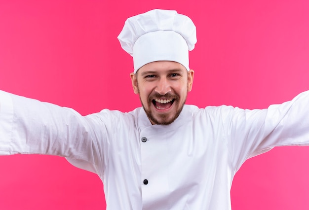 Gratis foto professionele mannelijke chef-kok in wit uniform en koken hoed wijd openende handen verwelkomend gebaar glimlachend vrolijk staande over roze achtergrond maken