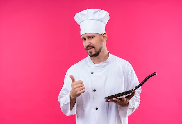 Professionele mannelijke chef-kok in wit uniform en kokhoed die een pan houdt die camera met zekere glimlach bekijkt die duimen toont die zich over roze achtergrond bevinden