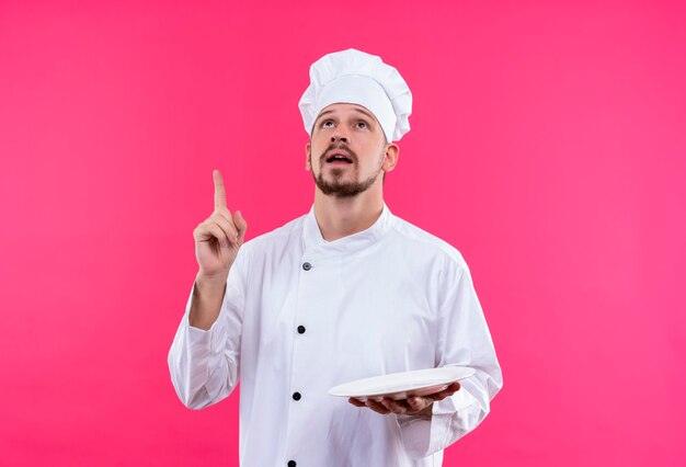 Professionele mannelijke chef-kok in wit uniform en kokhoed met een lege plaat die met vinger omhoog wijst gericht op een taak die zich over roze achtergrond bevindt