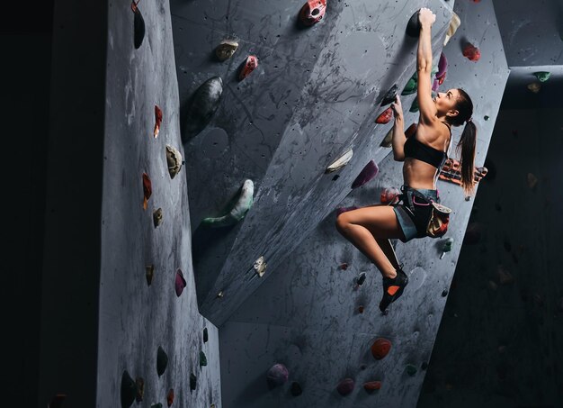 Professionele vrouwelijke klimmer die aan de bouldermuur hangt, oefen binnenshuis klimmen.