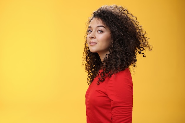 Profiel shot van stijlvolle en coole aantrekkelijke Europese 25-jarige vrouw met krullend haar in een rode jurk die naar de camera draait en breed glimlacht met opgetogen zorgeloze uitdrukking, plezier makend over gele achtergrond.