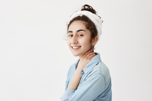 Profiel van een jong meisje met donker en golvend haar glimlachen, gekleed in denim shirt. Mooie vrouw met gelukkige glimlach, die en van het goede zonnige dag stellen tegen witte muur dromen genieten