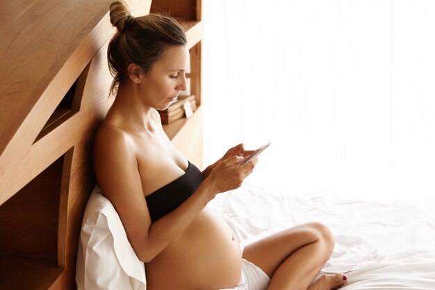 Profiel van ernstige zwangere vrouw met behulp van mobiele telefoon, bericht aan haar arts te typen, afspraak maken. Mooie vrouw verwacht baby met behulp van elektronisch apparaat, zittend op haar bed