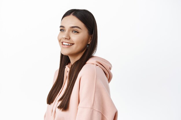 Profielopname van een glimlachende jonge vrouw die gelukkig kijkt naar de kopieerruimte aan de linkerkant, een roze hoodie draagt en tegen een witte muur staat