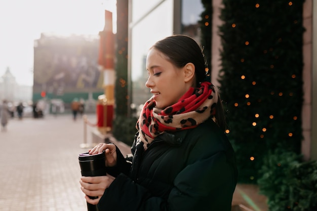 Profielportret van een brunette meisje met een warme donkere jas en sjaal geniet van koffie en loopt buiten in zonlicht en warme winterdag