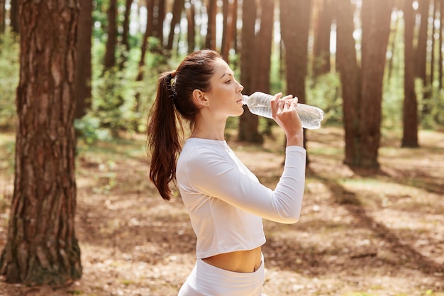 Profielportret van een mooie ontspannen atleet Europese vrouw in witte sportkleding die staat, rust, fles vasthoudt en drinkt terwijl ze wegkijkt