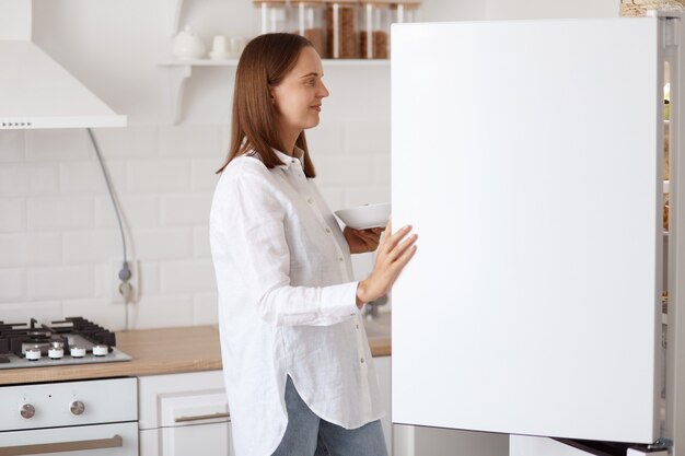 Profielportret van mooie jonge volwassen vrouw die een wit overhemd draagt, glimlachend in de koelkast met een aangename glimlach, een bord in handen, poserend met een keukenset op de achtergrond.
