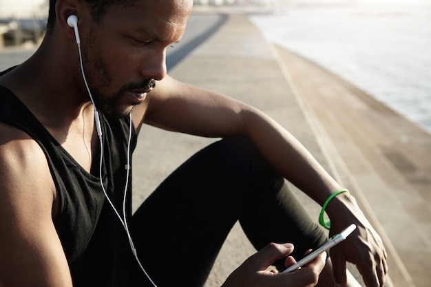 Profielportret van zwarte beambte in oortelefoons die rust hebben na zijn ochtend buitentraining, naar muziek luisteren, zijn smartphonescherm bekijken en favoriete liedjes uit de lijst met nummers kiezen