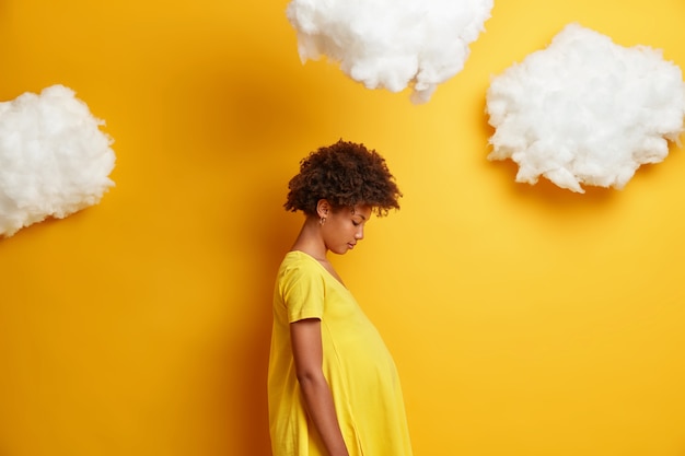 Profielschot van Afro-Amerikaanse vrouw met zwangere buik, kijkt neer op haar buik, draagt gele kleding voor zwangere vrouwen, heeft een grote buik, staat zijwaarts met wolken erboven, wacht op baby.