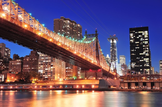 Queensboro Bridge over New York City East River bij zonsondergang met rivierreflecties en de skyline van Midtown Manhattan verlicht.