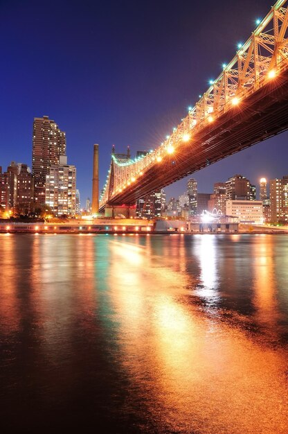 Queensboro Bridge over New York City East River bij zonsondergang met rivierreflecties en de skyline van Midtown Manhattan verlicht.