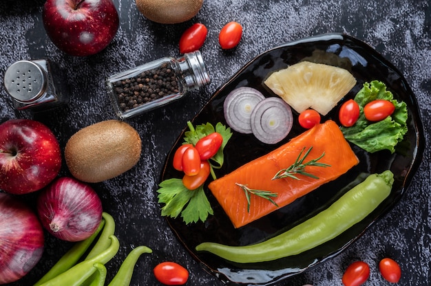 Rauwe zalmfilets, peper, kiwi, ananas en rozemarijn op een bord en zwarte cementvloer.