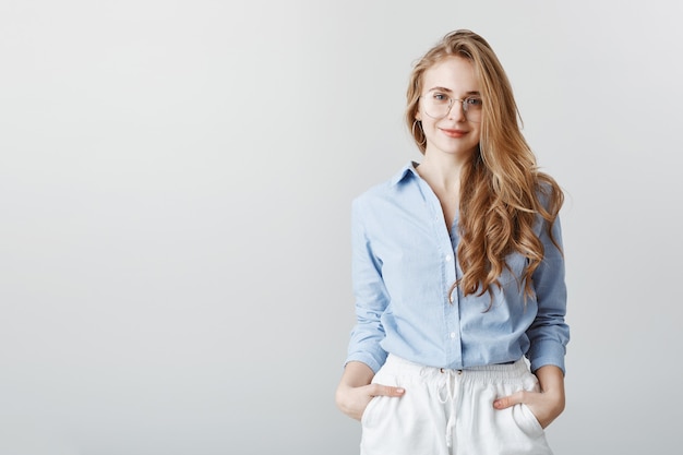 Reisagent hoort de wensen van de klant. Portret van knappe europese zakenvrouw in blauwe blouse en bril, hand in hand in de zakken en glimlachend, vriendelijk en beleefd over grijze muur