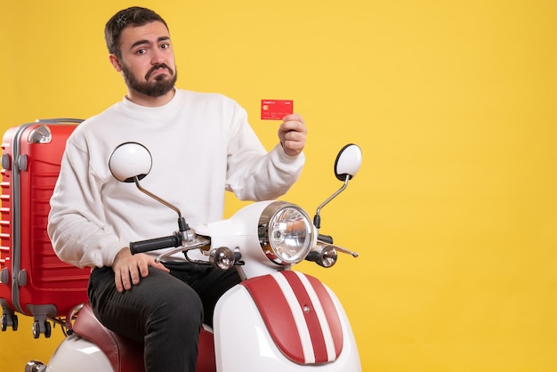 Reisconcept met een jonge verwarde reizende man die op een motorfiets zit met een koffer erop met een bankkaart op geel